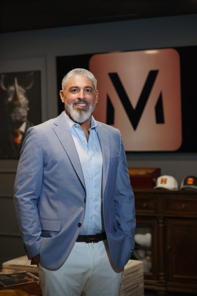 Smiling man in a light-blue blazer and white pants stands indoors, backdrop features a pink logo panel with a bold black symbol.